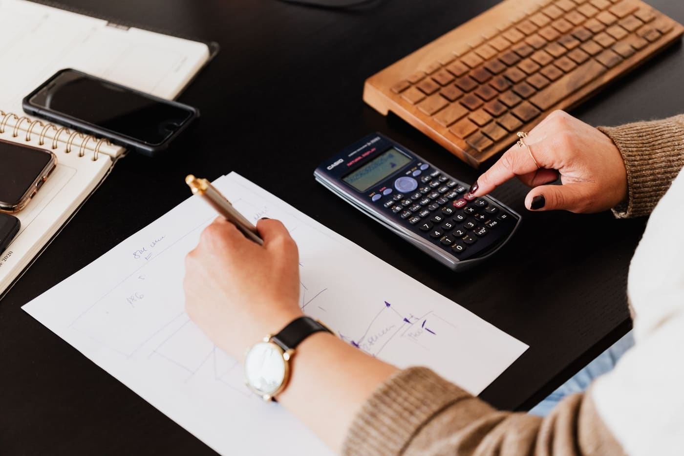 A hand on a calculator beside handwritten notes and a wooden keyboard on a dark desk