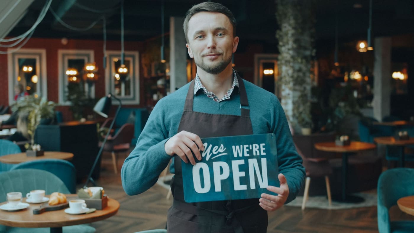 A cafe owner in an apron holding a we are open sign inside the cafe