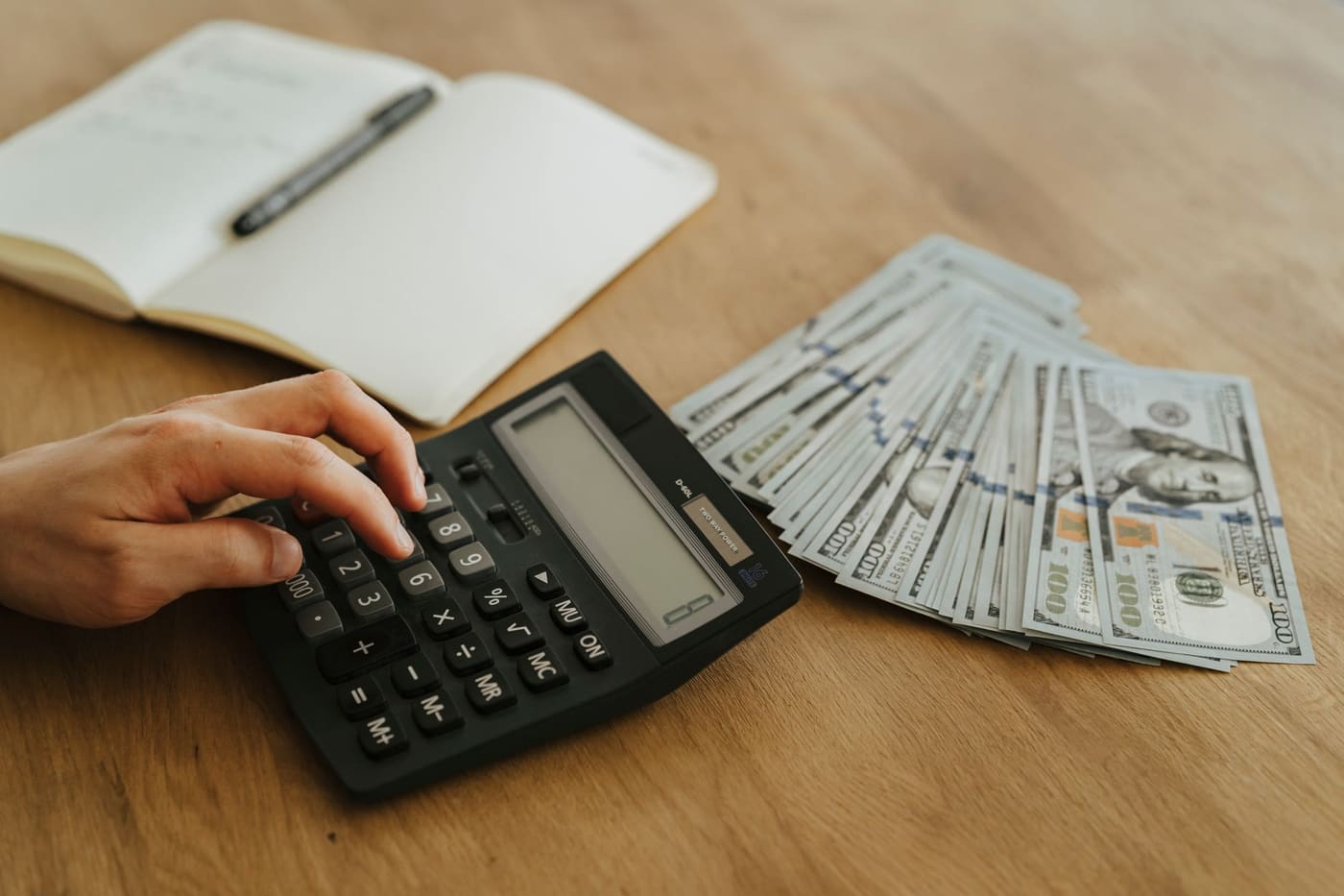 A hand using a calculator beside a notebook and a stack of banknotes on a wooden table