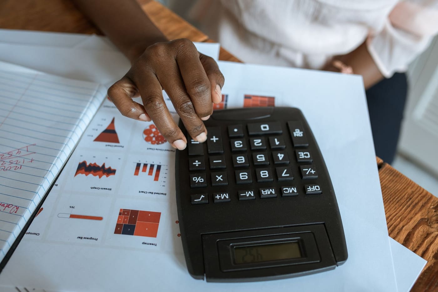 Hands pressing a black calculator on a desk covered with printed charts