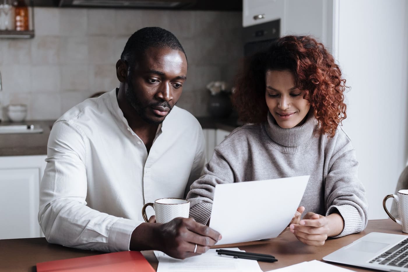 A couple sit at a kitchen table reading a document together with a laptop and coffee