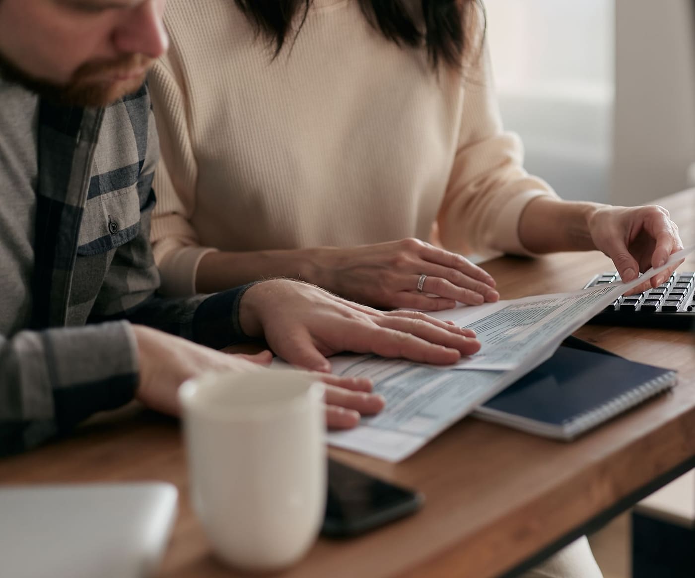 Two people's hands on printed financial papers at a wooden table with a keyboard and mug nearby