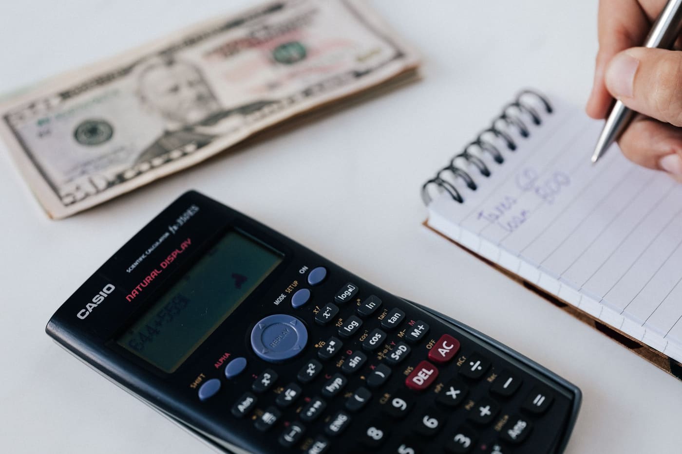 A notebook, scientific calculator and banknotes seen from above with a pen writing