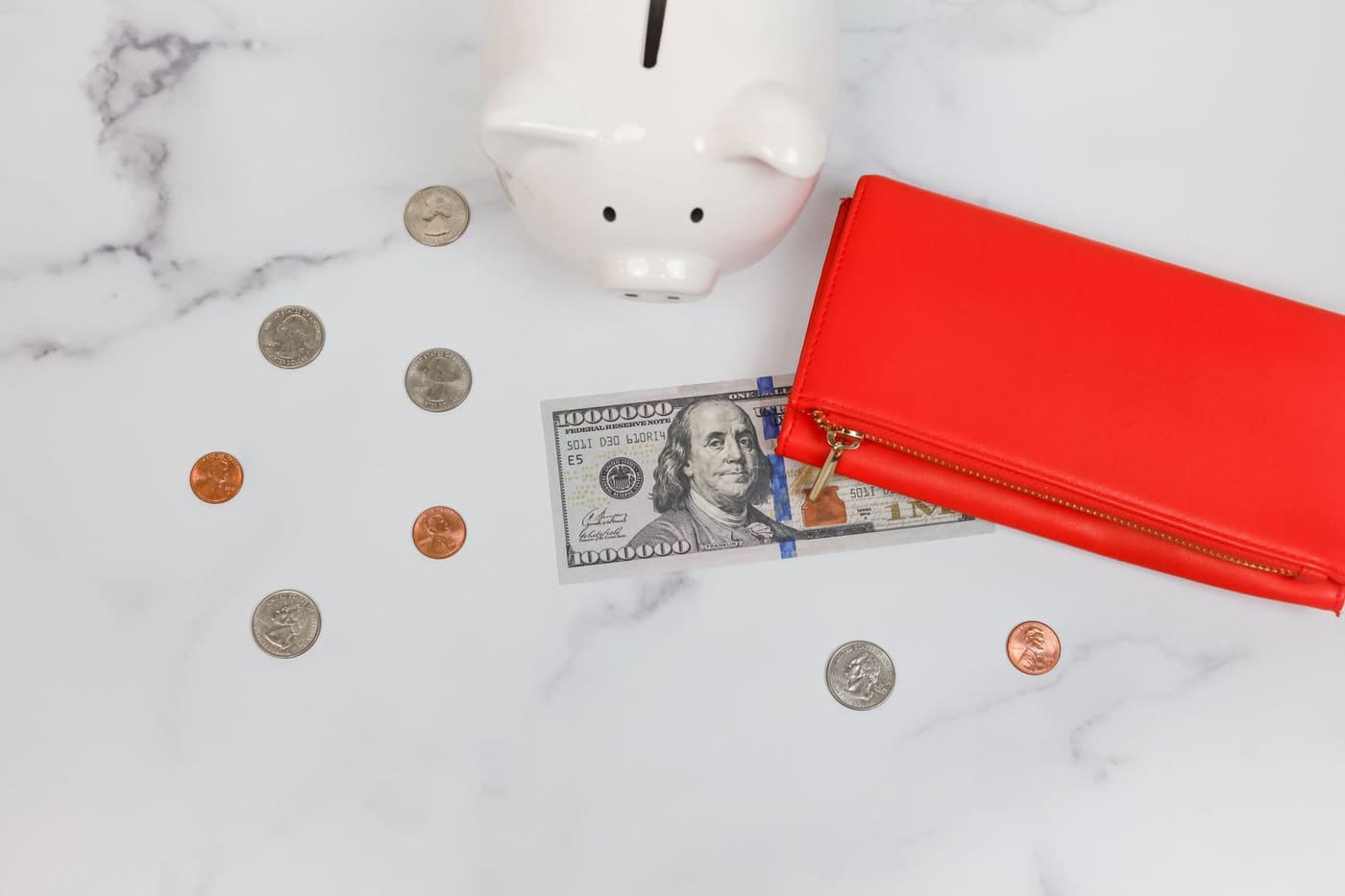 A white piggy bank, scattered coins, a bill and a red wallet on a marble surface
