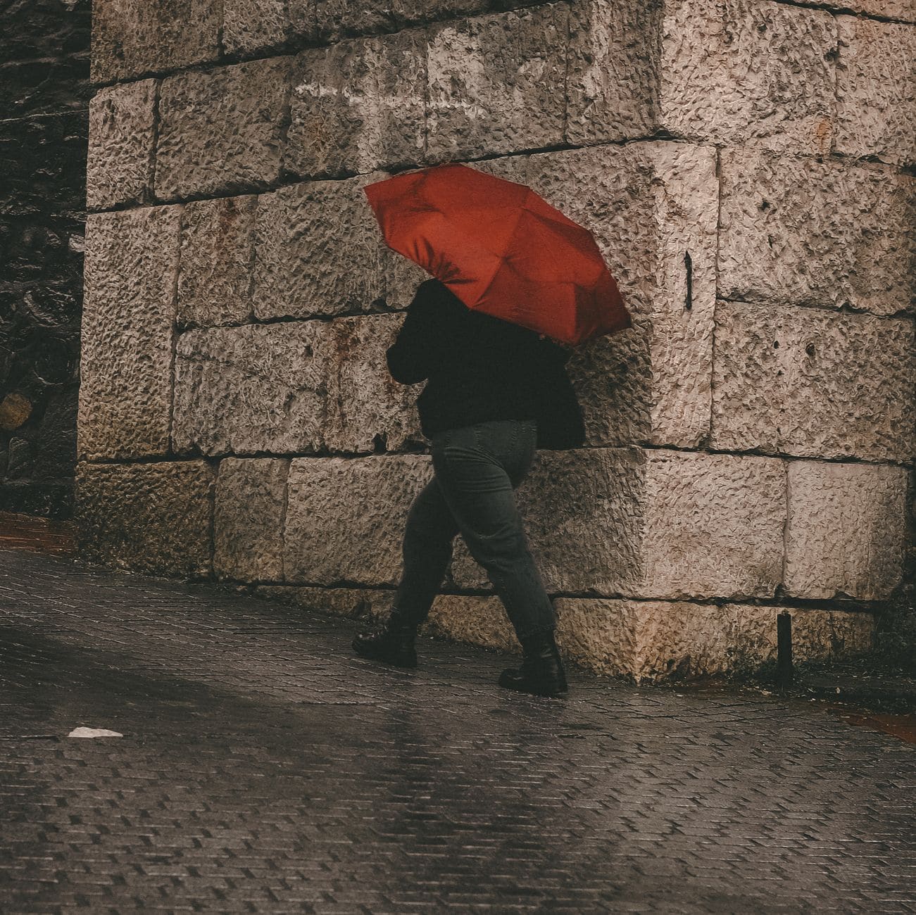 A person with a red umbrella walking past a stone wall in the rain